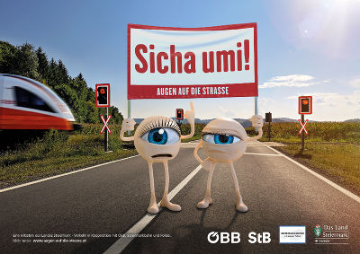 Auf dem Bild ist ein Zug zu sehen, daneben stehen zwei Maskottchen, die ein Plakat hochhalten, worauf steht: Sicha umi